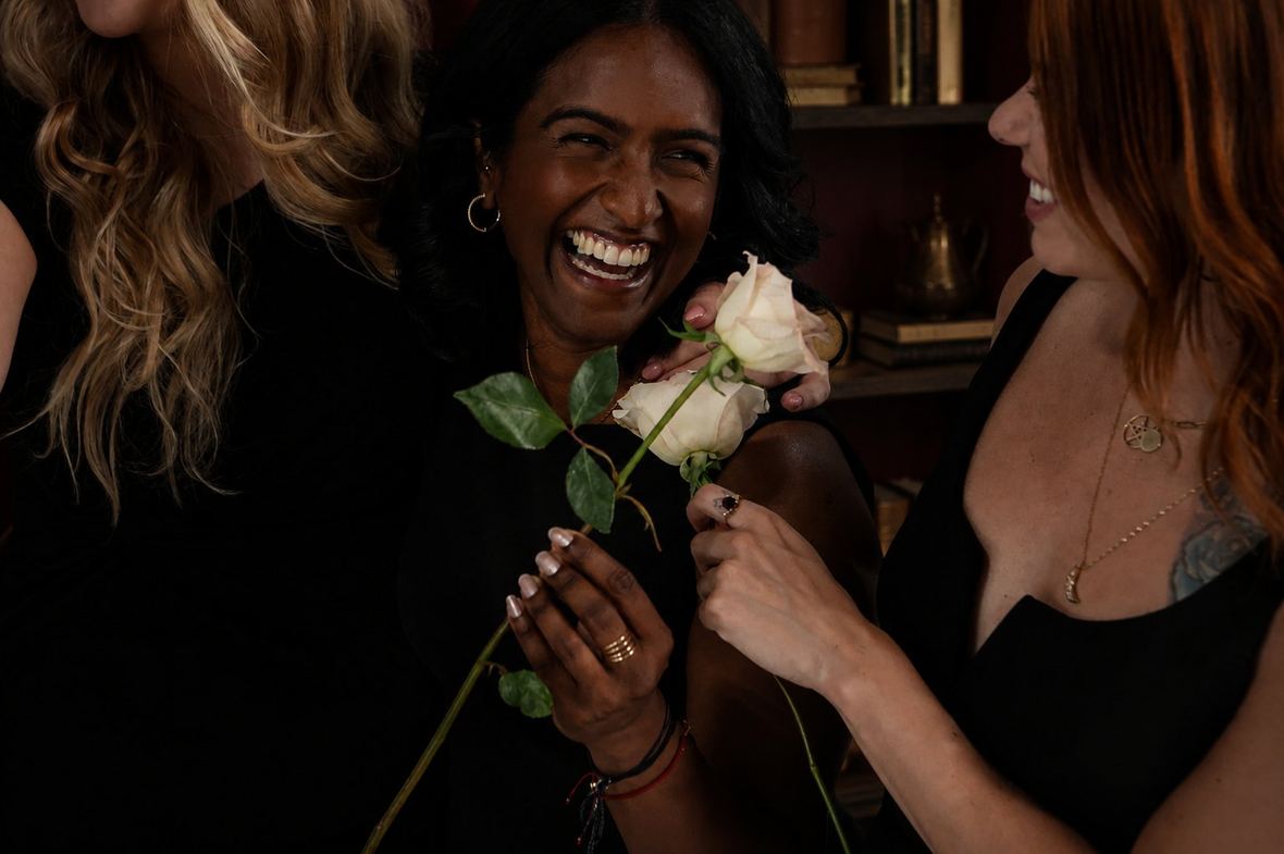 Women laughing together, holding white roses, in a deep red room